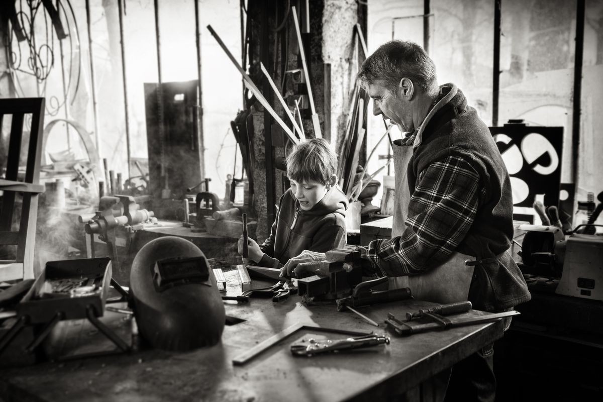 A craftsman in his craft workshop teaches his young son how to work on metal pieces. Black and white